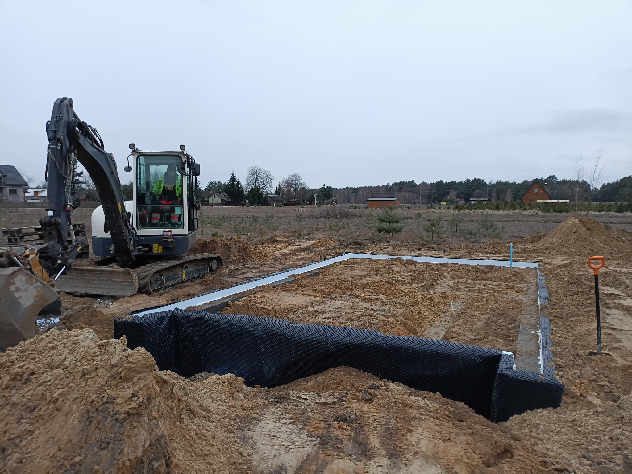 Mini koparka przy wykopie pod fundamenty z izolacją pionową. Widoczny pracownik w kabinie i ułożona folia izolacyjna. W tle zabudowa jednorodzinna i pochmurne niebo.