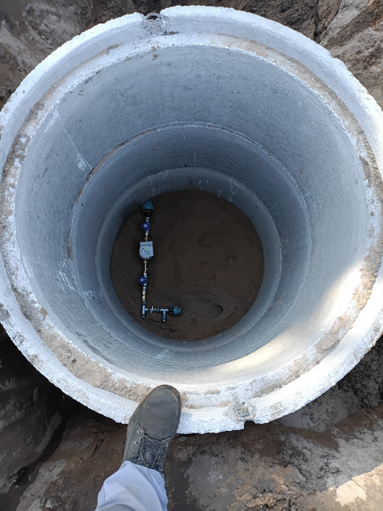 Betonowy krąg szamba z zainstalowaną armaturą wodną, widok z góry. But robotnika w rogu kadru sugeruje skalę. Świeżo wykonana instalacja wewnątrz.