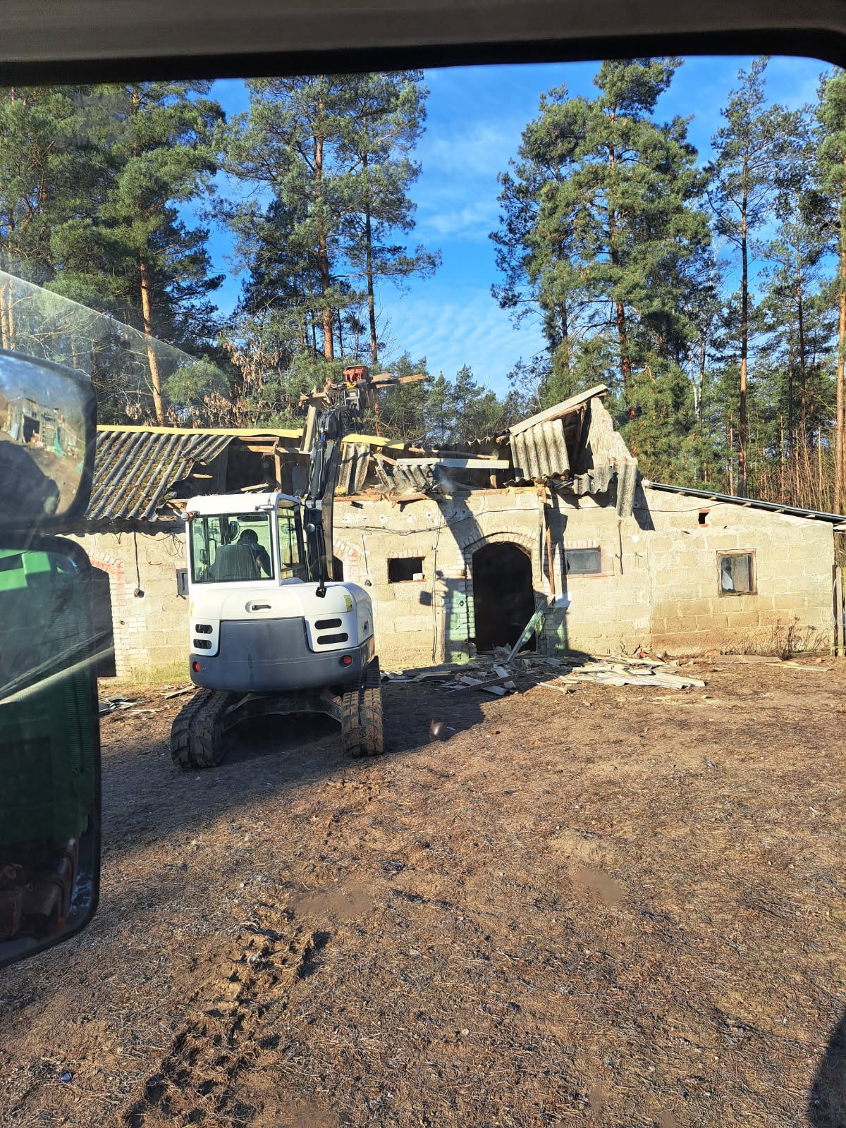 Mini koparka gąsienicowa w trakcie rozbiórki starego, parterowego budynku z cegły. Widoczne uszkodzenia dachu i ścian. W tle las iglasty i błękitne niebo.