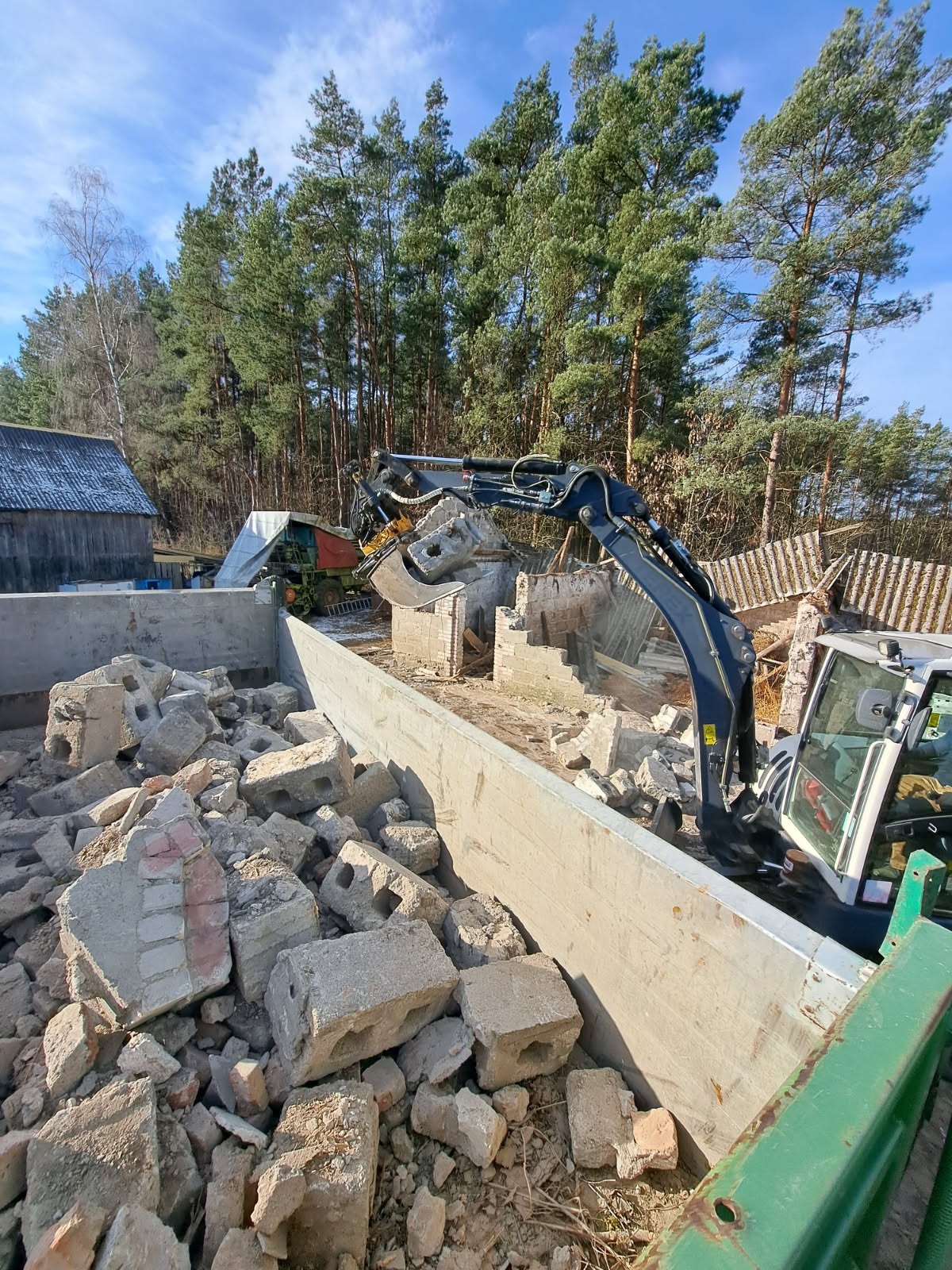 Mini koparka z łyżką pełną gruzu na tle rozbieranego budynku i lasu. Widoczny fragment kontenera na gruz. Ujęcie z góry, w tle budynek gospodarczy.