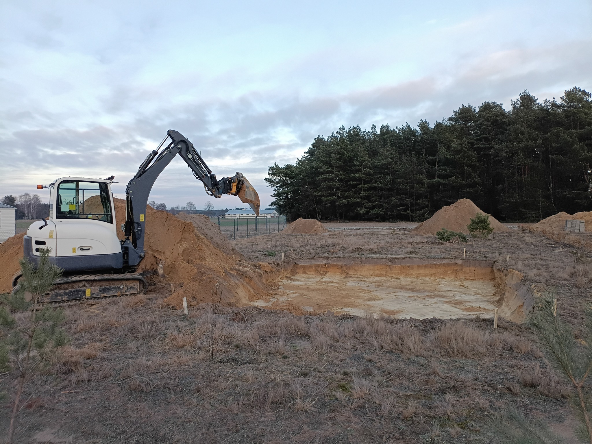 Biała mini koparka przy wykopie pod fundamenty na działce z usypaną ziemią i lasem w tle, pochmurne niebo.
