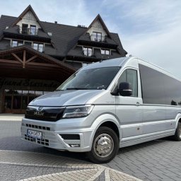 NikoTour - Nowoczesny, srebrny bus MAN z przyciemnianymi szybami, zaparkowany na brukowanym podjeździe przed hotelem Grand Stasinda o charakterystycznej góralskiej architekturze.
