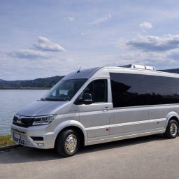 NikoTour - Srebrny bus MAN TGE z przyciemnianymi szybami i dachową klimatyzacją, zaparkowany na asfalcie przy jeziorze, z zielonymi wzgórzami w tle.