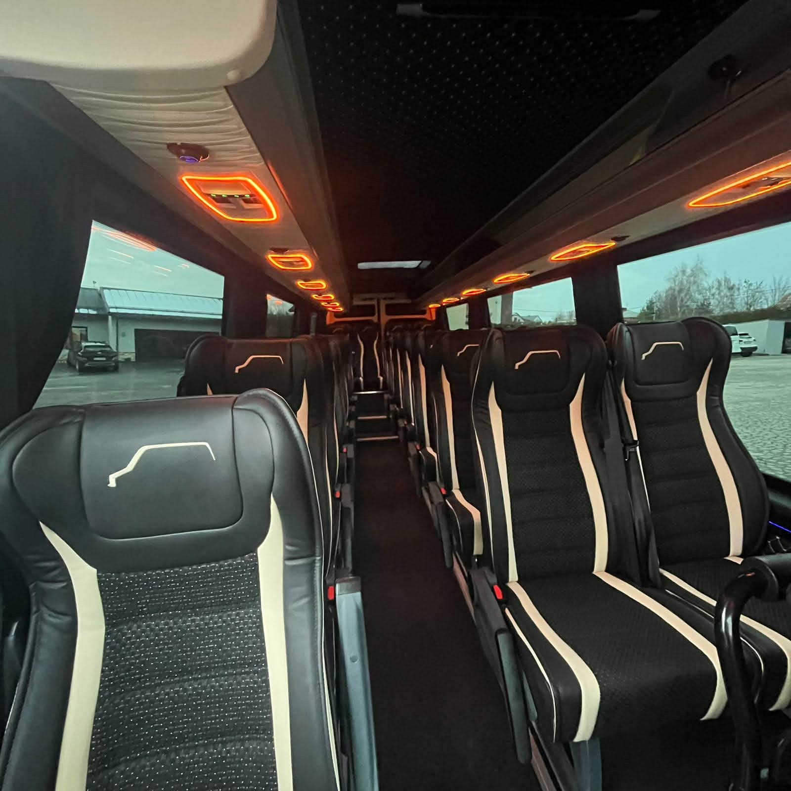 Wnętrze luksusowego busa z czarnymi skórzanymi fotelami z beżowymi wstawkami i logo. Oświetlenie LED wzdłuż sufitu. Widok na zewnątrz przez okna.
