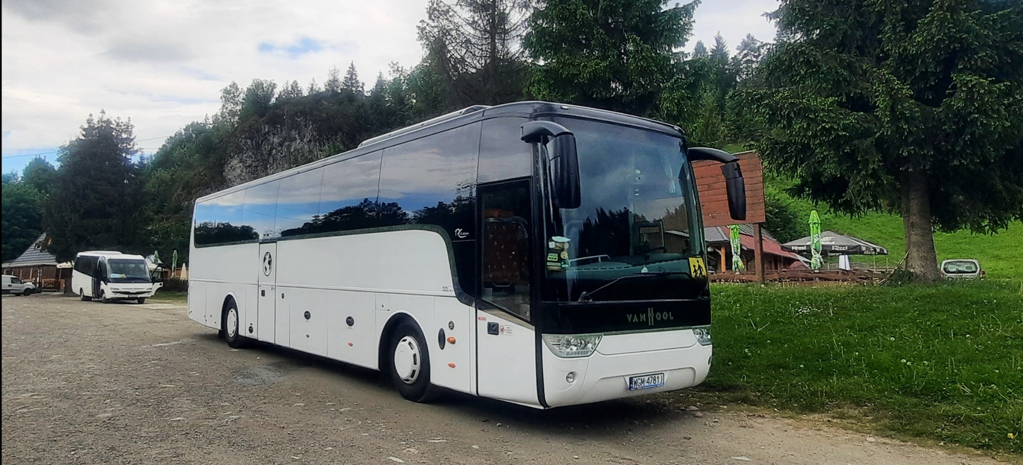 Biały autokar Van Hool na żwirowym parkingu, z widocznym fragmentem drugiego autokaru w tle i zielonym krajobrazem górskim. Tablica rejestracyjna WGM 4781J.