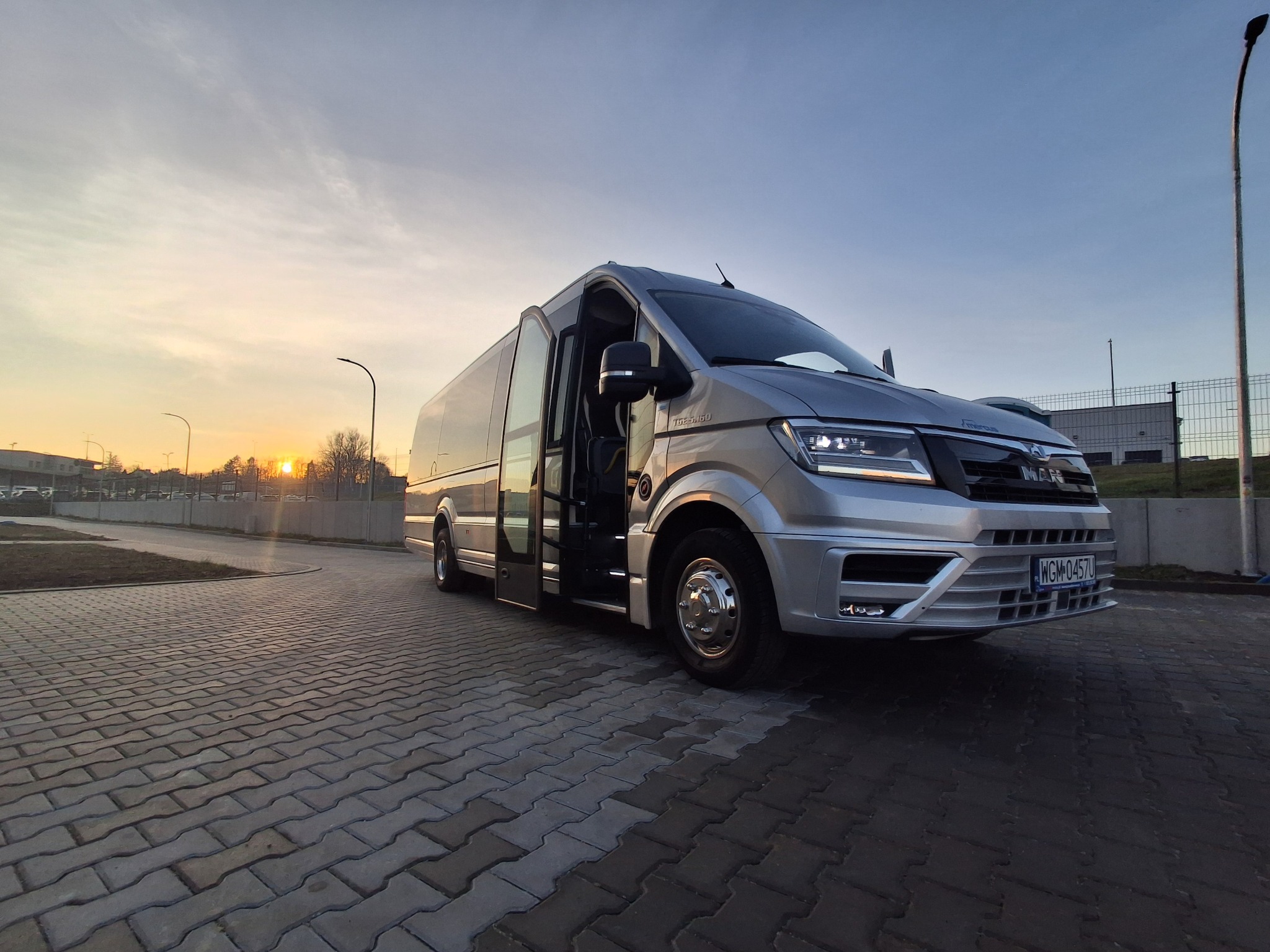 Srebrny bus MAN z otwartymi drzwiami na brukowanym placu, w tle zachodzące słońce. Widok z niskiej perspektywy, podkreślający nowoczesny design pojazdu.