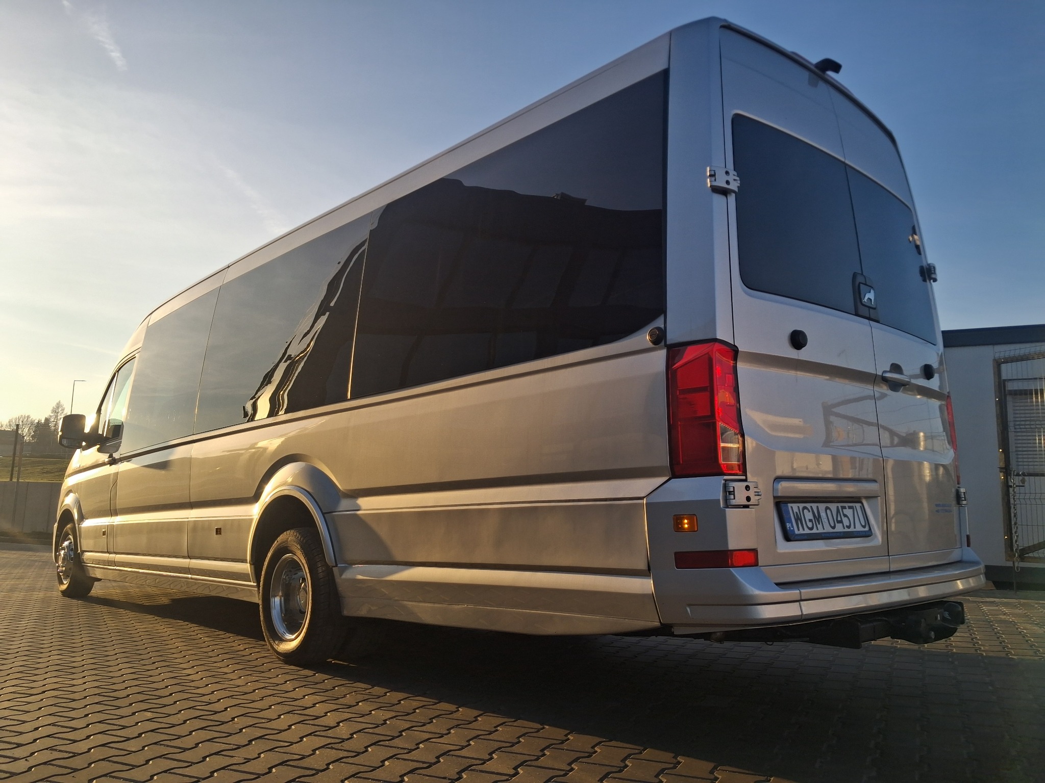 Srebrny bus z przyciemnianymi szybami, widziany z tyłu, zaparkowany na kostce brukowej w słoneczny dzień. Widoczna tablica rejestracyjna i hak holowniczy.