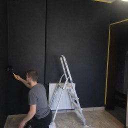 D.M. Daniel Maciejowski - Mężczyzna maluje ścianę na czarno wałkiem, klęcząc na podłodze zabezpieczonej folią. Widoczna drabina, grzejnik i taśma malarska przy suficie. Oświetlenie sufitowe w rogu.