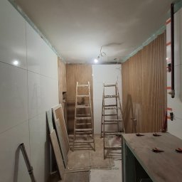 D.M. Daniel Maciejowski - Wnętrze w trakcie prac wykończeniowych: białe płytki na ścianie, drewniane panele, dwie drabiny, narzędzia glazurnicze. Widoczne elementy instalacji.