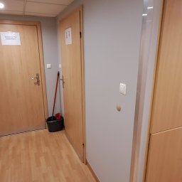 D.M. Daniel Maciejowski - Korytarz biurowy z drewnianą podłogą i szarą ścianą. Widoczne drzwi z oznaczeniami toalet. Wiadro i mop stoją przy ścianie. Oświetlenie sufitowe.