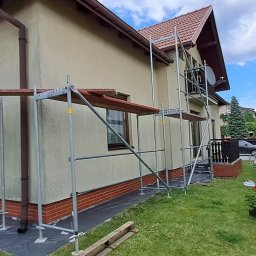 D.M. Daniel Maciejowski - Dom jednorodzinny w trakcie prac elewacyjnych, otoczony rusztowaniem. Widoczne zabezpieczenia podłoża. Ujęcie z poziomu trawnika, w tle zieleń i błękitne niebo.