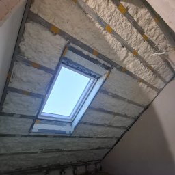 D.M. Daniel Maciejowski - Izolacja poddasza pianą PUR z widoczną konstrukcją metalową i oknem dachowym. Biała piana wypełnia przestrzeń między profilami. Widok na skośny sufit w trakcie prac.