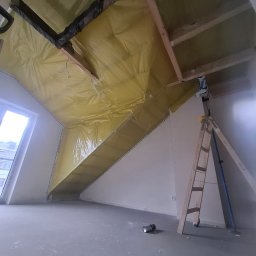 D.M. Daniel Maciejowski - Poddasze w trakcie izolacji: żółta folia paroizolacyjna na skośnym suficie, drabina oparta o ścianę, surowa podłoga. Widoczne drewniane elementy konstrukcyjne.