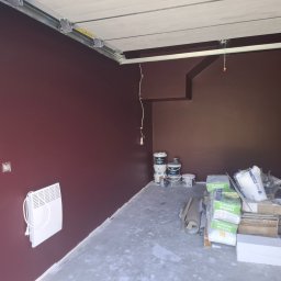 D.M. Daniel Maciejowski - Pomieszczenie po malowaniu ścian na bordowo, widoczny grzejnik, puszki z farbą i materiały budowlane. Szara podłoga i biały sufit.