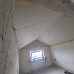 D.M. Daniel Maciejowski - Pokój na poddaszu w trakcie prac wykończeniowych: ściany z surowym tynkiem, zamontowana płyta karton-gips na skosach dachu, okno. Widok z dołu, surowy stan.