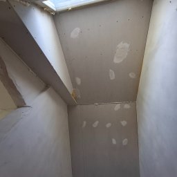 D.M. Daniel Maciejowski - Poddasze z oknem dachowym w trakcie wykańczania płytami gipsowo-kartonowymi. Widoczne łączenia płyt i miejsca po szpachlowaniu. Ujęcie z dołu, podkreślające skosy.