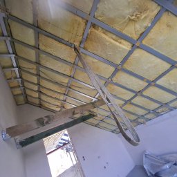 D.M. Daniel Maciejowski - Montaż sufitu podwieszanego z izolacją z wełny mineralnej, widoczne profile metalowe i fragment ściany z elementami konstrukcyjnymi. Ujęcie z dołu.