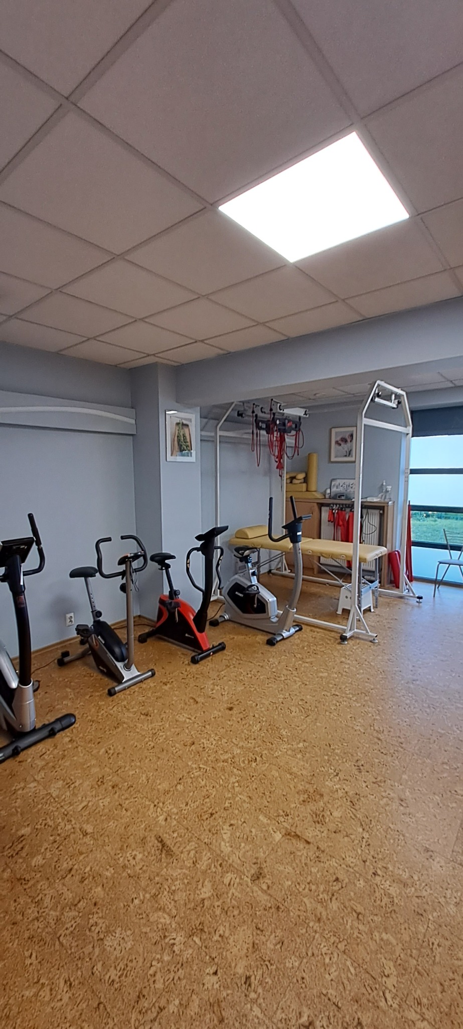 Jasne wnętrze sali rehabilitacyjnej z czterema rowerami stacjonarnymi i stołem do masażu. Podłoga korkowa, ściany szare, sufit kasetonowy z oświetleniem.