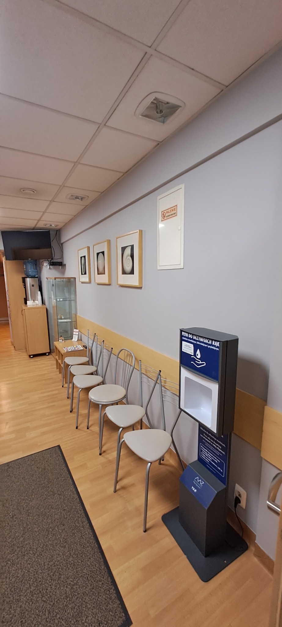 Jasne wnętrze poczekalni z rzędem krzeseł, dozownikiem płynu do dezynfekcji rąk i dekoracjami ściennymi. Widoczne elementy wyposażenia biura.