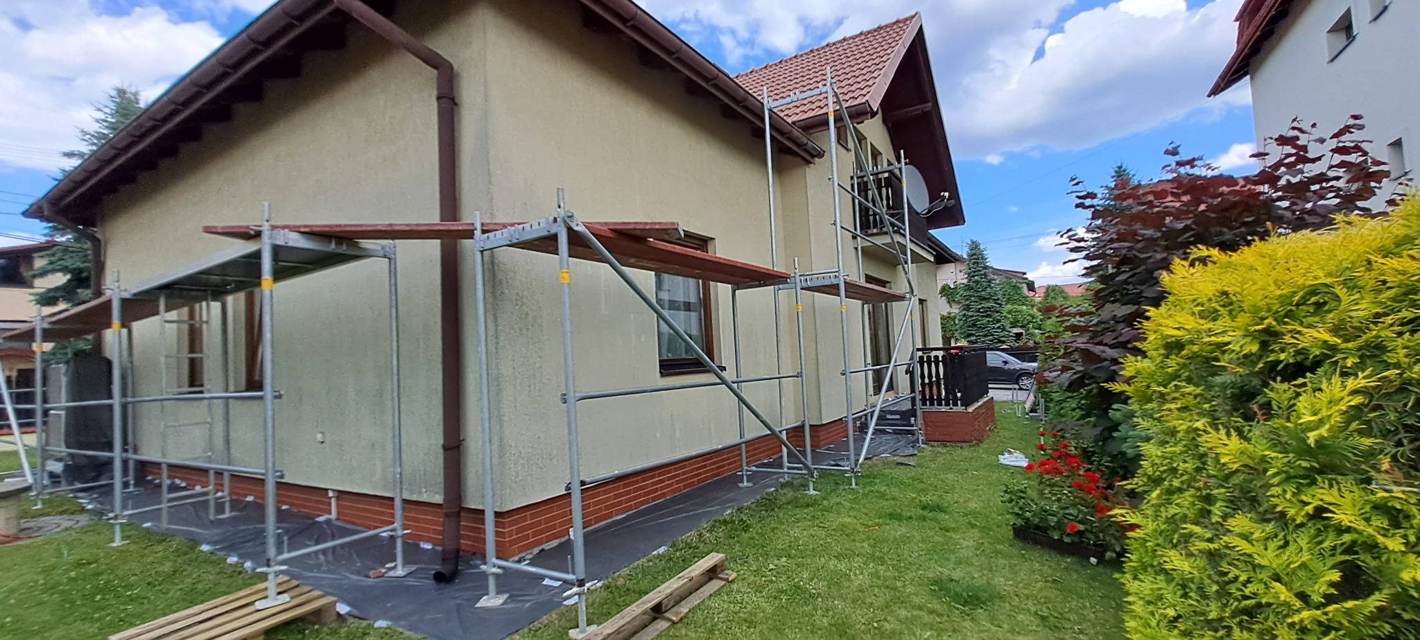 Dom jednorodzinny w trakcie prac elewacyjnych, otoczony rusztowaniem. Widoczne zabezpieczenia podłoża. Ujęcie z poziomu trawnika, w tle zieleń i błękitne niebo.