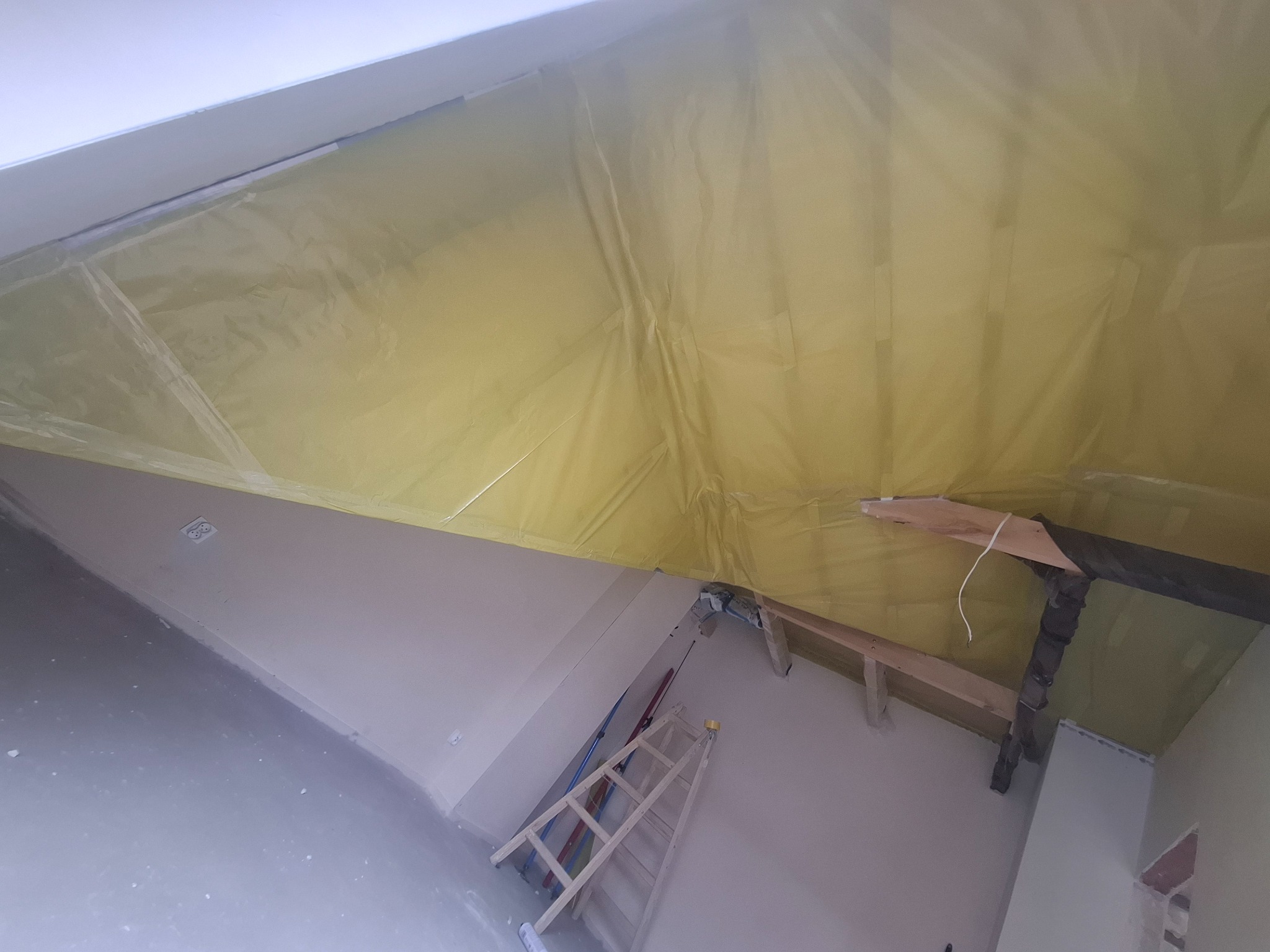 Skośny sufit zabezpieczony folią malarską, widoczne drewniane elementy konstrukcyjne i drabina oparta o ścianę. Przygotowanie do prac wykończeniowych na poddaszu.