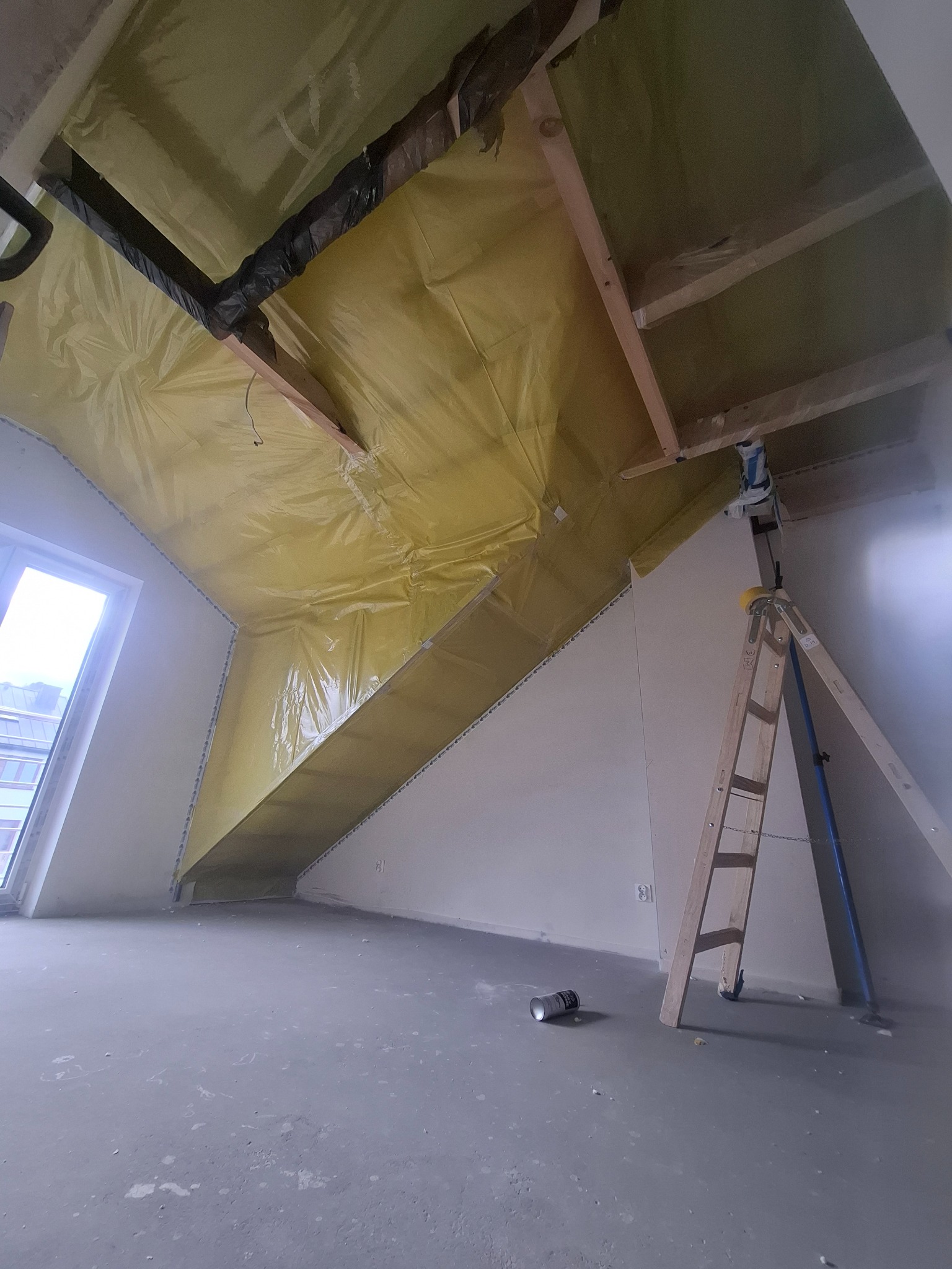 Poddasze w trakcie izolacji: żółta folia paroizolacyjna na skośnym suficie, drabina oparta o ścianę, surowa podłoga. Widoczne drewniane elementy konstrukcyjne.