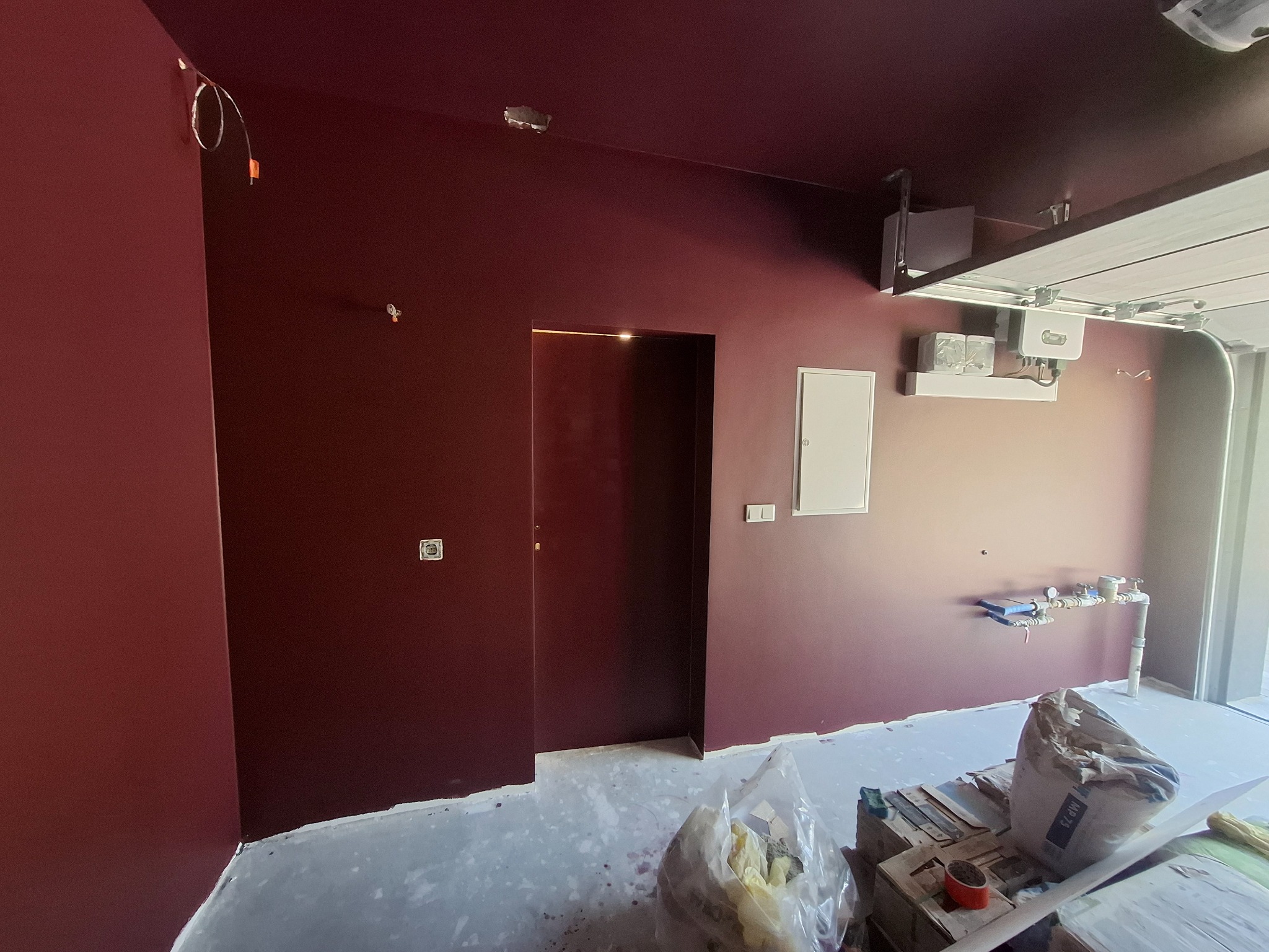 Pomieszczenie po malowaniu ścian i sufitu na bordowo. Widoczne niedociągnięcia przy podłodze i niezabezpieczone kable. W tle brama garażowa.