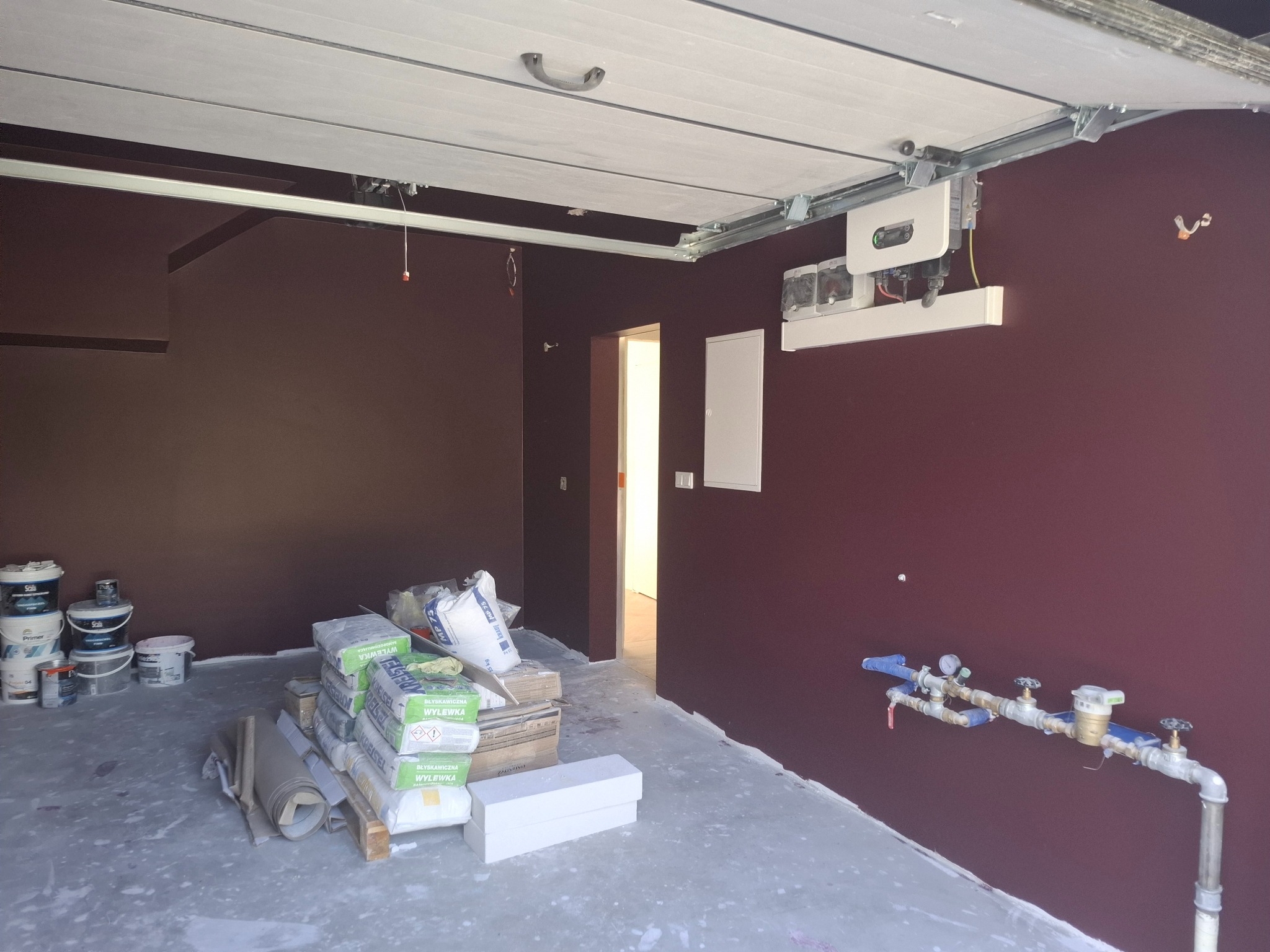 Wnętrze w trakcie prac wykończeniowych: pomalowane ściany w odcieniach bordo, materiały budowlane na podłodze, instalacja wodna przy ścianie, sufit z elementami konstrukcyjnymi.