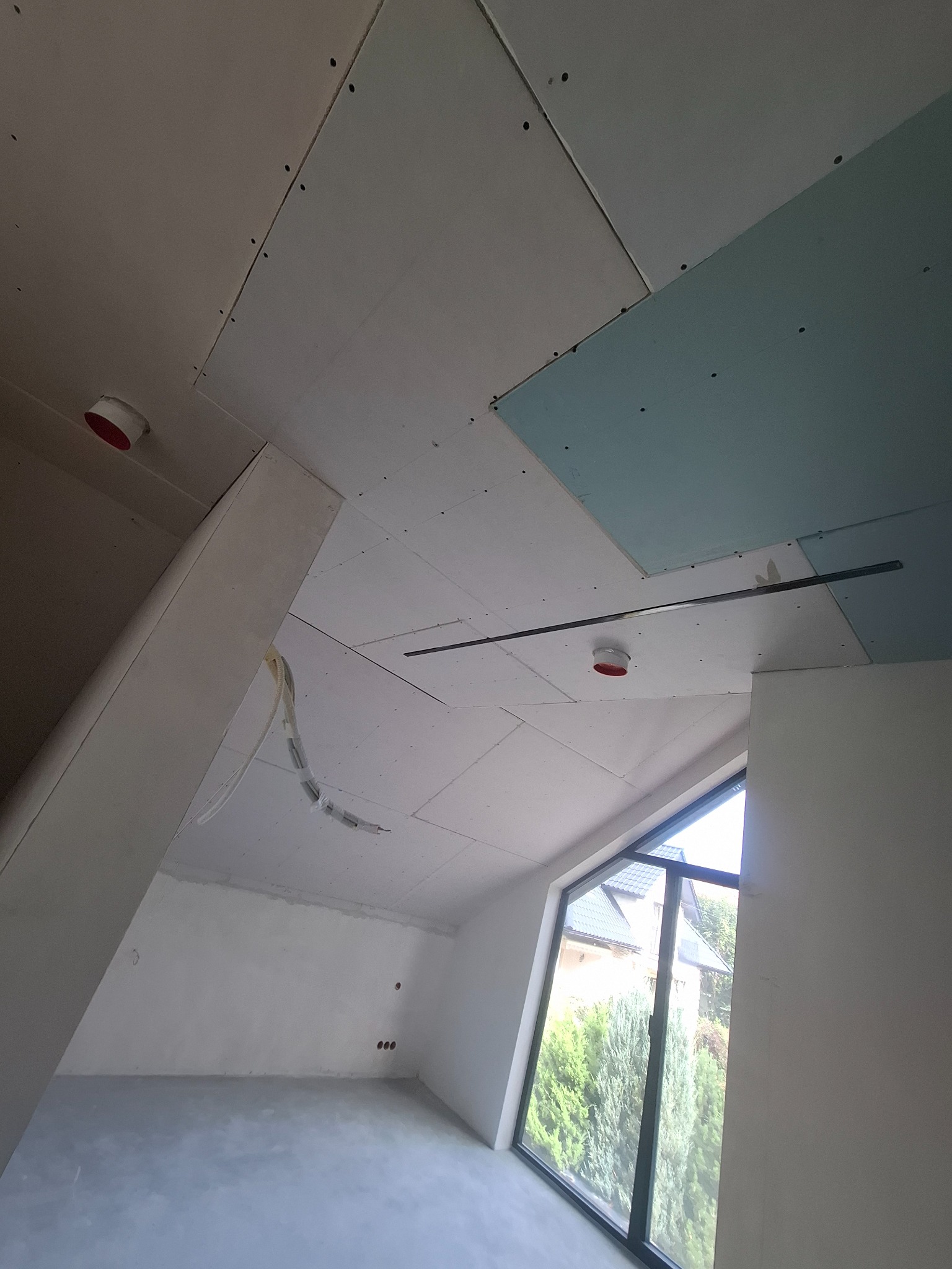 Skośny pokój na poddaszu w trakcie montażu płyt gipsowo-kartonowych na suficie, widoczne kable i okno z widokiem na zieleń.