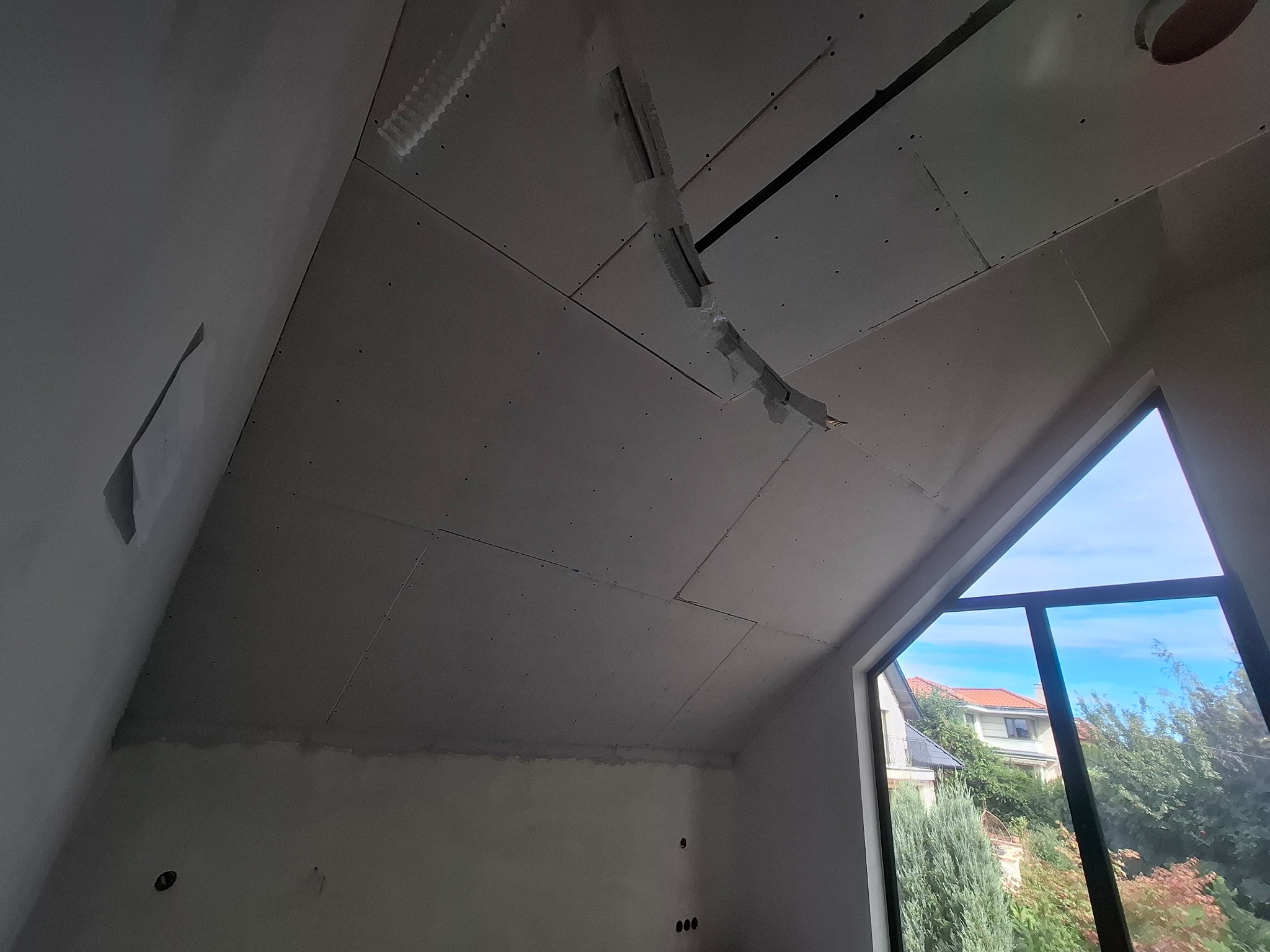 Skośny sufit z płyt kartonowo-gipsowych w trakcie montażu, widoczne łączenia i elementy mocujące. W tle okno z widokiem na zieleń.