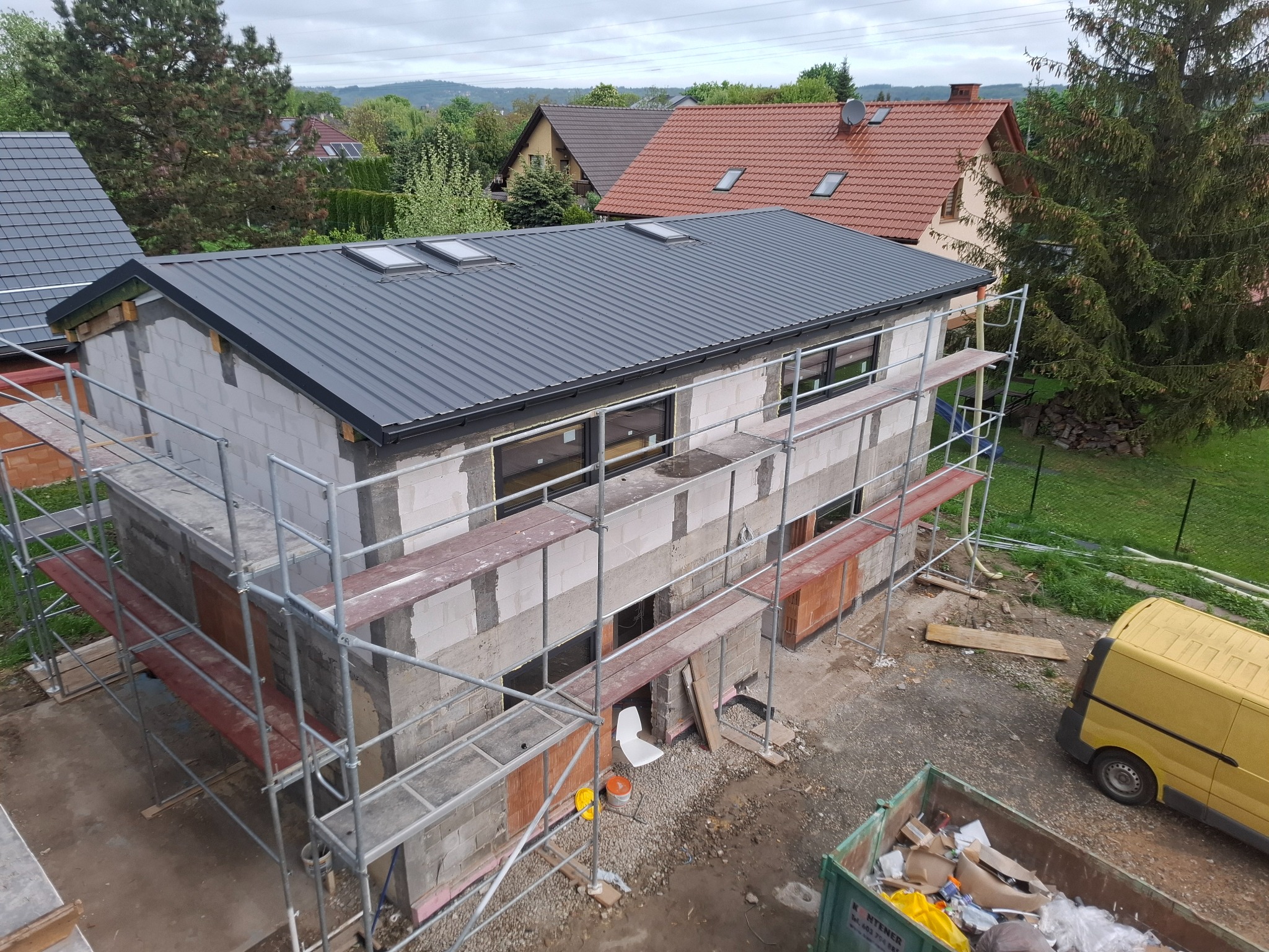 Budowa domu z szarej blachy na dachu, widok z góry, rusztowania, okna wstawione, śmietnik i żółty bus w tle, etap surowy zamknięty.