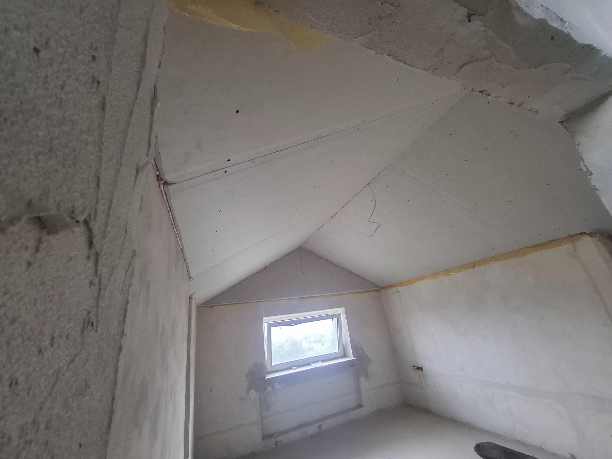 Pokój na poddaszu w trakcie prac wykończeniowych: ściany z surowym tynkiem, zamontowana płyta karton-gips na skosach dachu, okno. Widok z dołu, surowy stan.