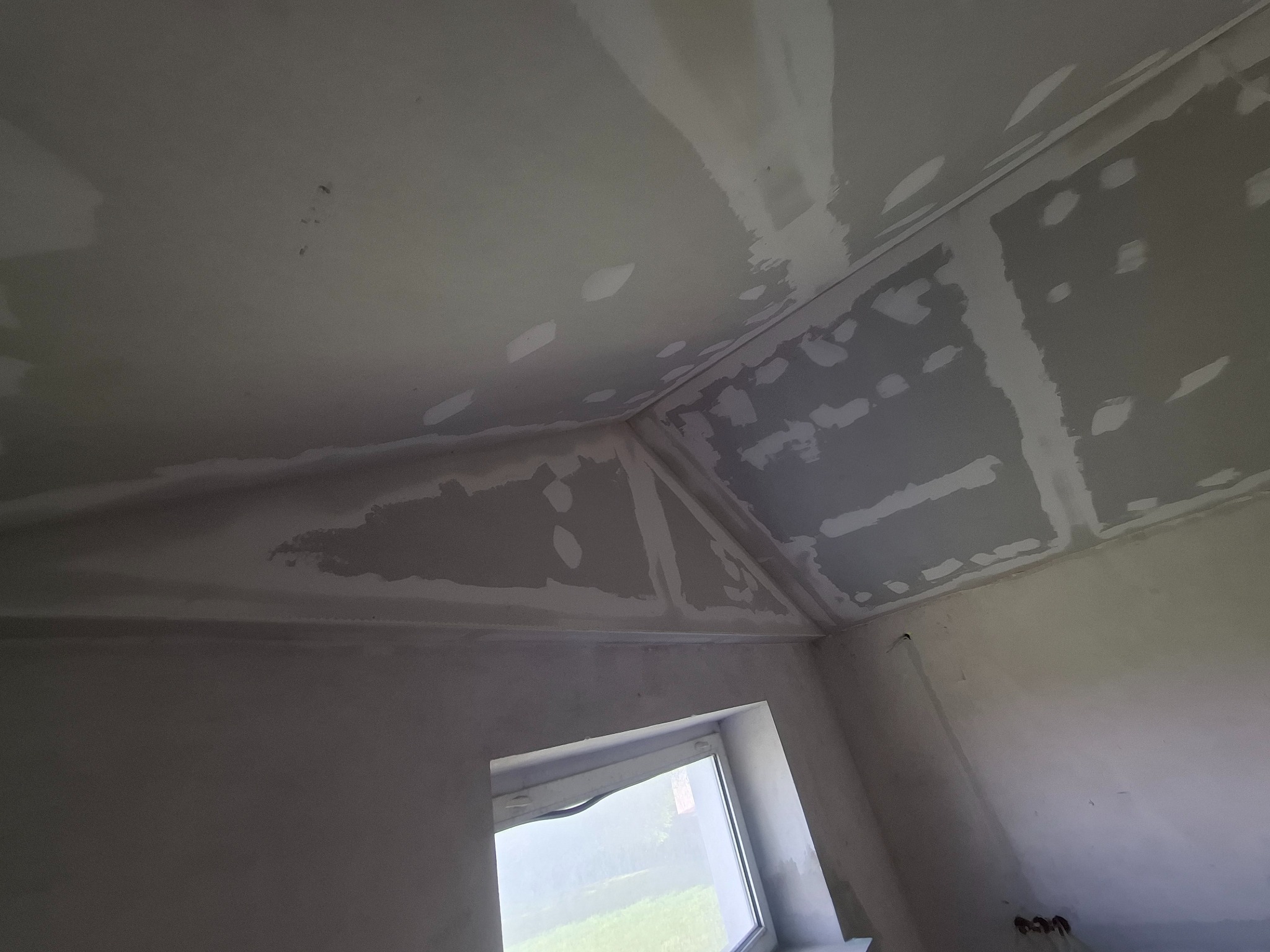 Skośny sufit poddasza po szpachlowaniu, widoczne łączenia płyt gipsowych i miejsca po wkrętach. W tle okno z widokiem na zieleń. Ściany przygotowane do malowania.