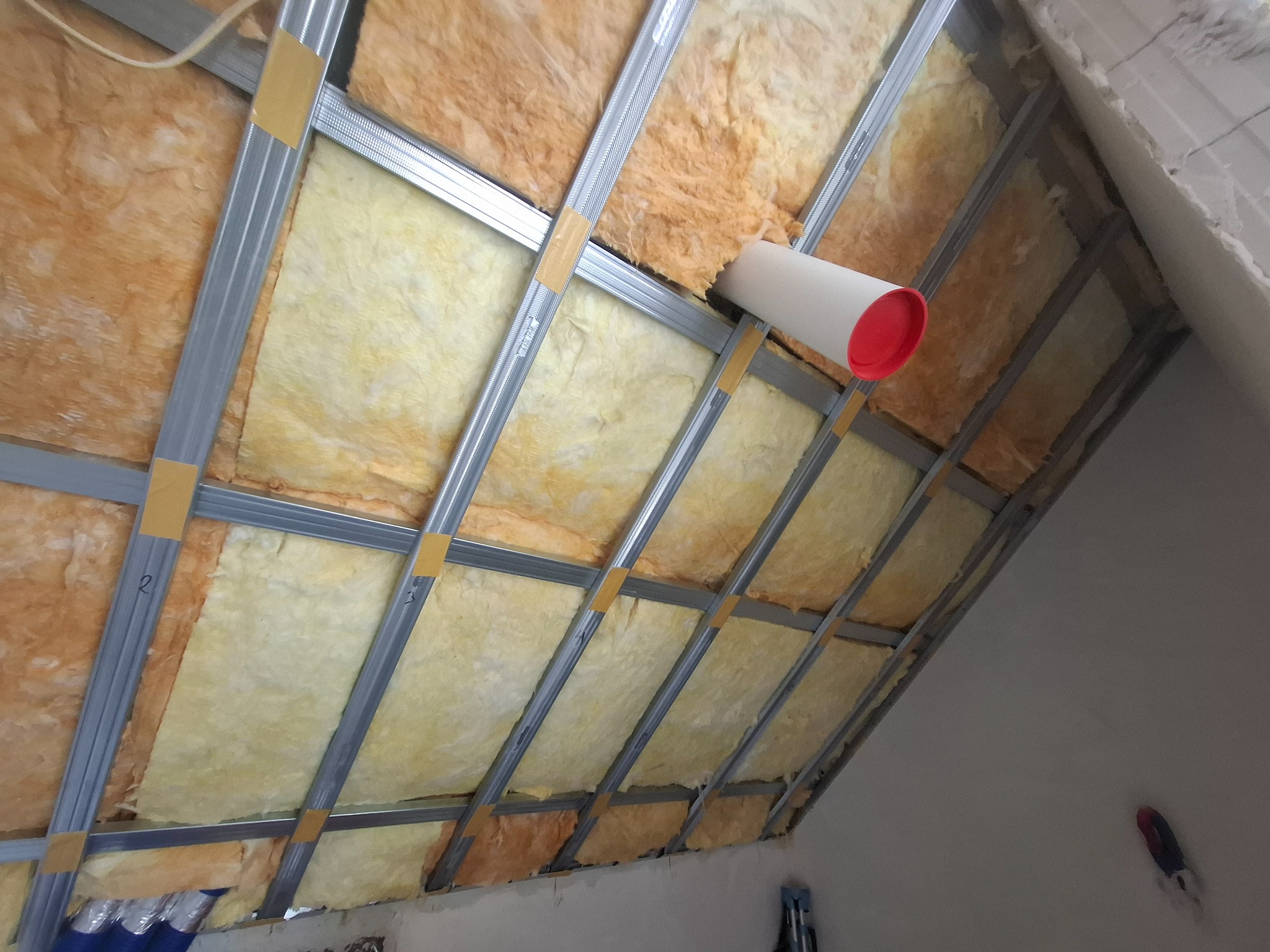 Skośny sufit poddasza z izolacją z wełny mineralnej, zamocowaną do metalowej konstrukcji. Widoczny fragment wentylacji. Ujęcie z dołu, w trakcie prac.