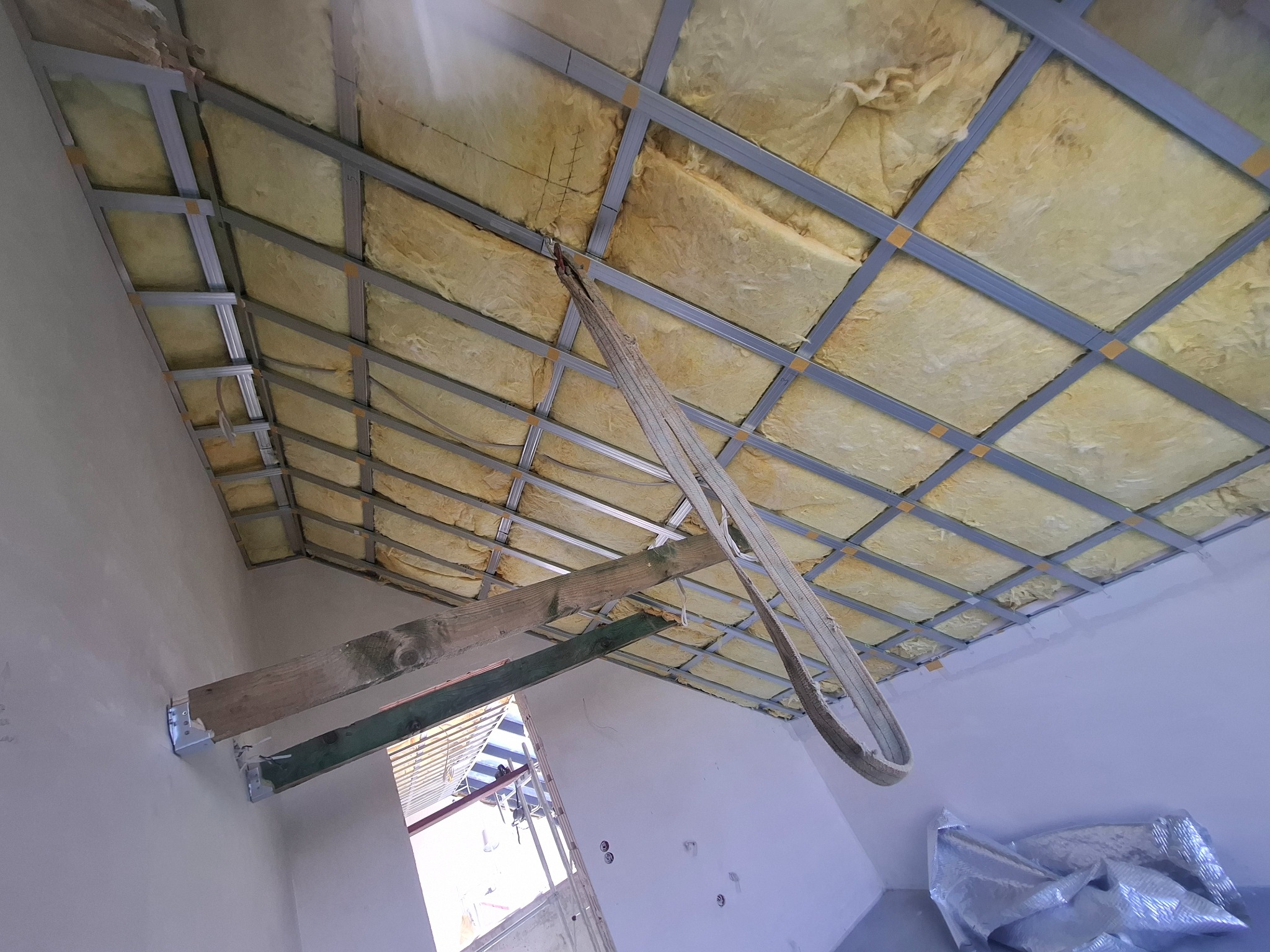 Montaż sufitu podwieszanego z izolacją z wełny mineralnej, widoczne profile metalowe i fragment ściany z elementami konstrukcyjnymi. Ujęcie z dołu.