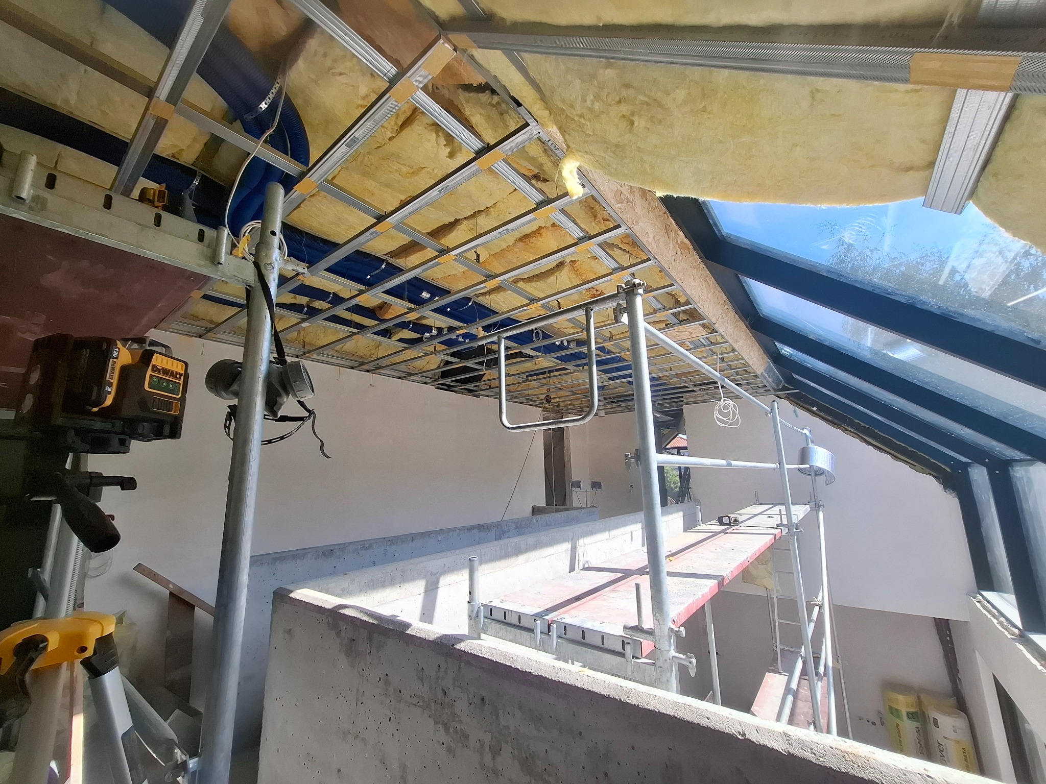 Montaż izolacji poddasza w Krzczonowie. Widoczna konstrukcja metalowa, wełna mineralna, rusztowanie i poziomica laserowa. Prace wykończeniowe w trakcie.