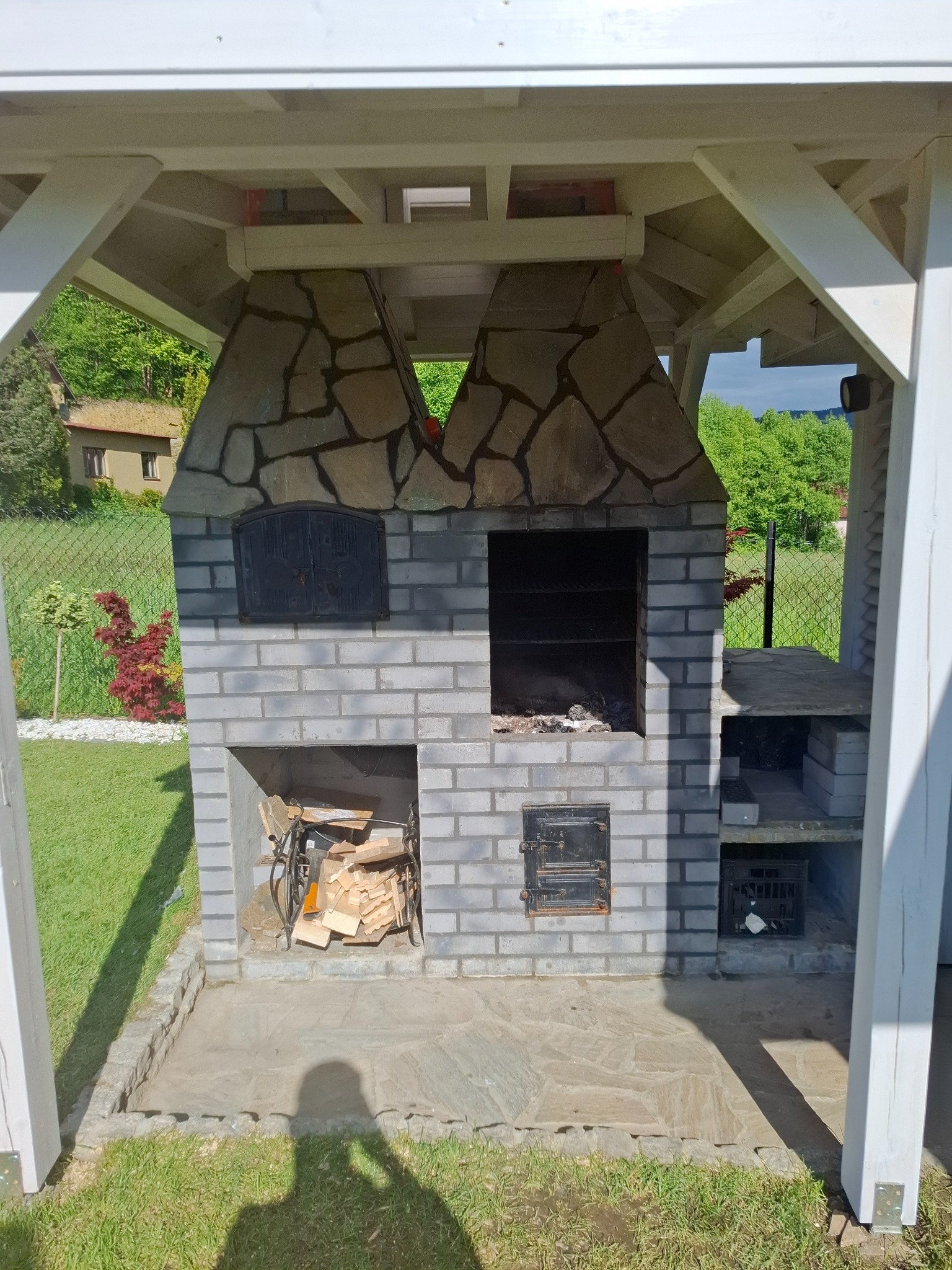 Murowany grill ogrodowy z szarej cegły i kamiennym wykończeniem pod zadaszeniem, z miejscem na drewno i akcesoria. Widok z przodu, oświetlenie dzienne.