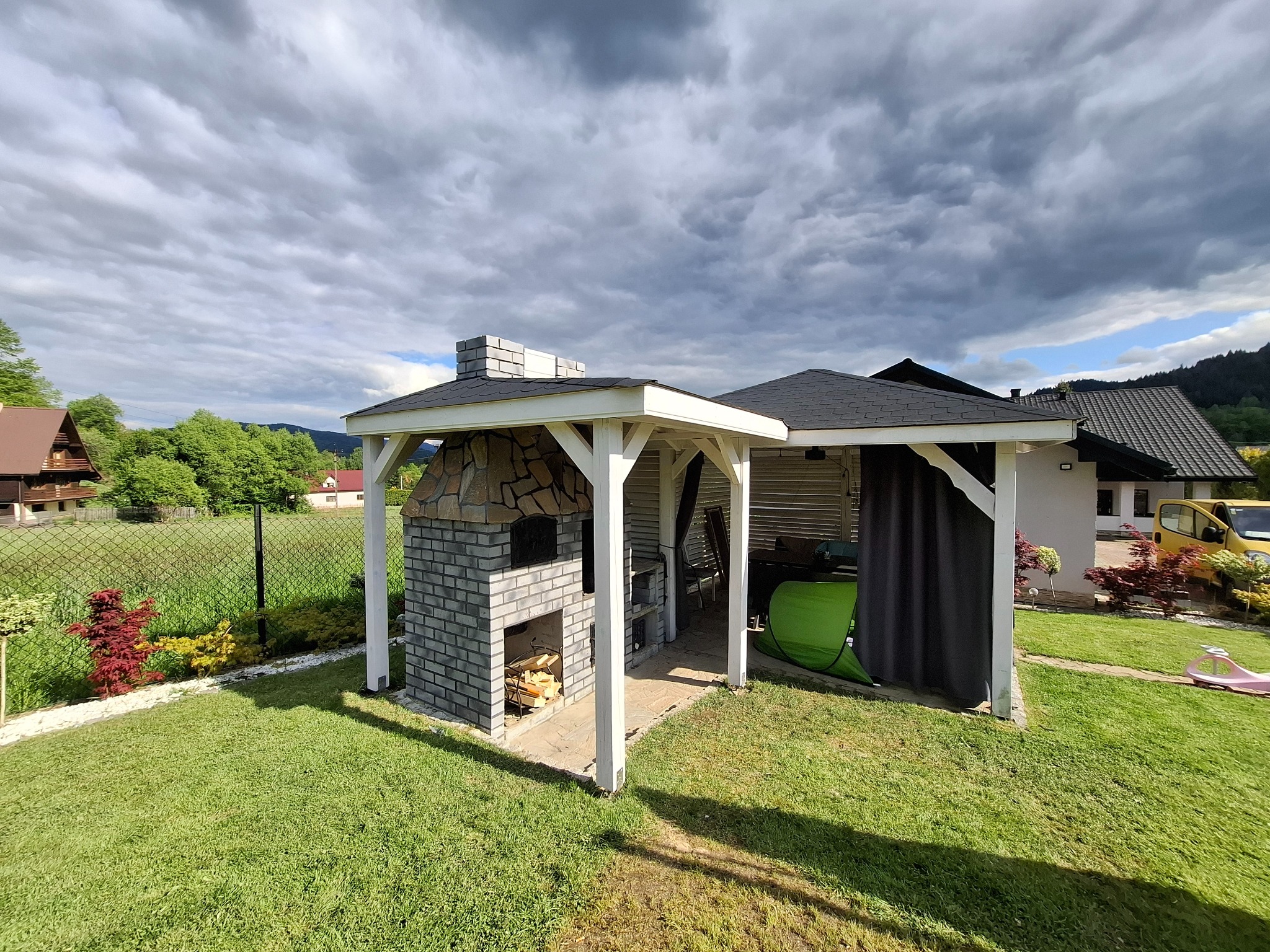 Murowany grill z kamiennymi elementami pod zadaszeniem na trawniku, z widocznym drewnem w palenisku. W tle domy i zieleń, pochmurne niebo.