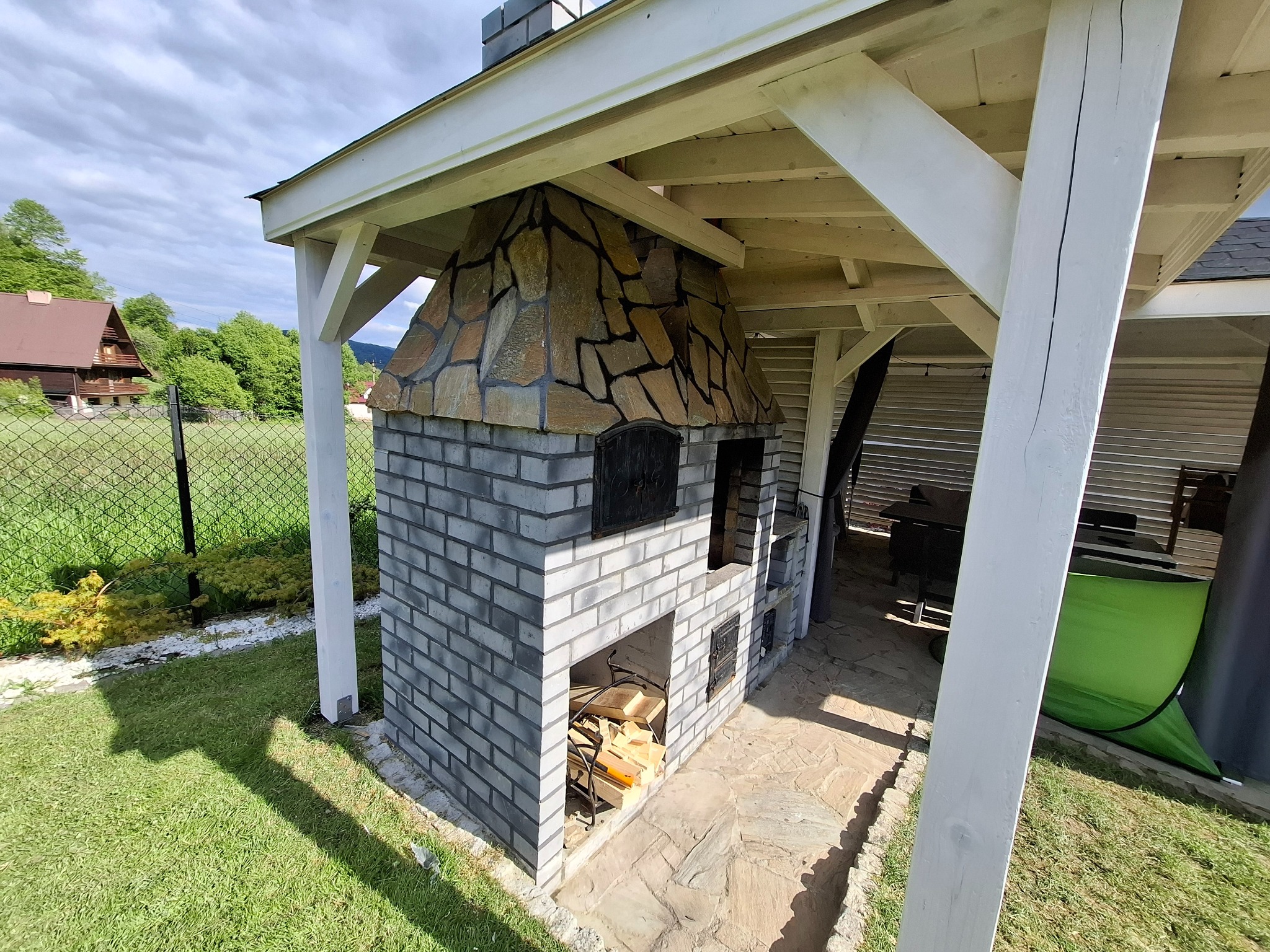 Murowany grill z kamienną obudową pod drewnianą wiatą, z zapasem drewna w dolnej części. Widok na ogród i dom w tle, w słoneczny dzień.