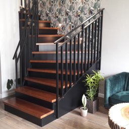 Estica Produkcja i Usługi Stolarskie - Nowoczesne schody z drewnianymi stopniami i czarną metalową balustradą, na tle tapety w tropikalne liście. Obok zielona sofa i stolik z plastra drewna.
