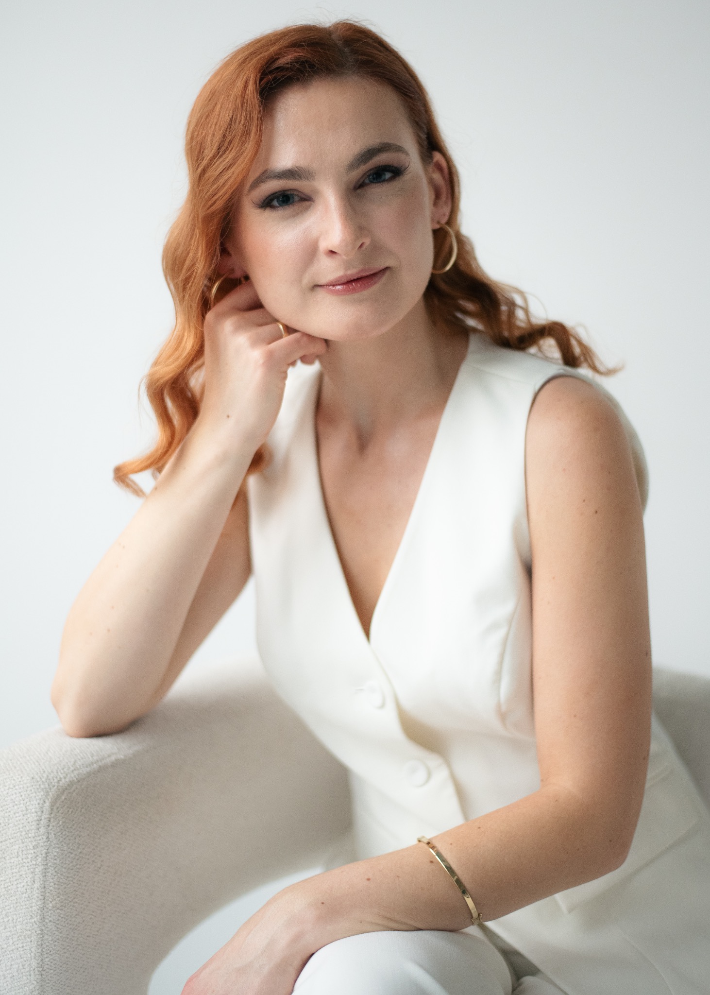 Portret kobiety z rudymi włosami, ubranej w białą kamizelkę, siedzącej na jasnym fotelu. Delikatny uśmiech, złota bransoletka i kolczyki. Neutralne tło.