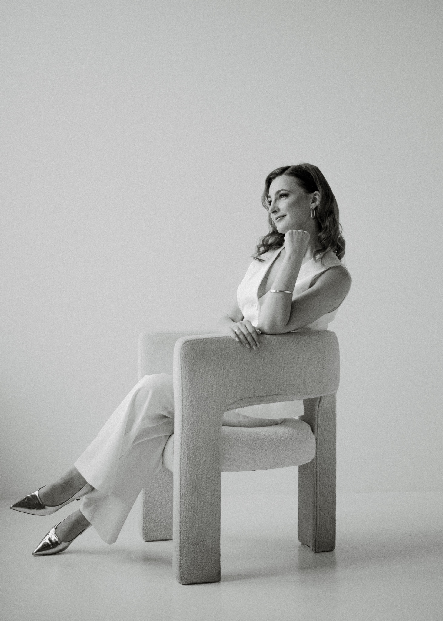 Portret kobiety w białym garniturze siedzącej na fotelu w minimalistycznym studio. Uśmiechnięta, zamyślona, patrzy w górę. Srebrne szpilki dodają elegancji.