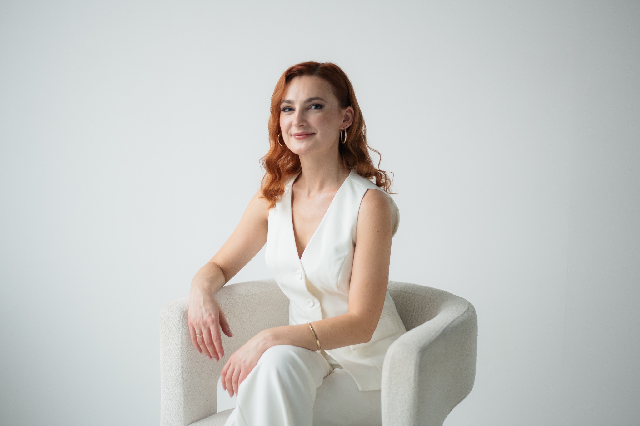 Portret kobiety z rudymi włosami w białym garniturze siedzącej w fotelu na jasnym tle. Uśmiechnięta, z biżuterią. Profesjonalny wizerunek.