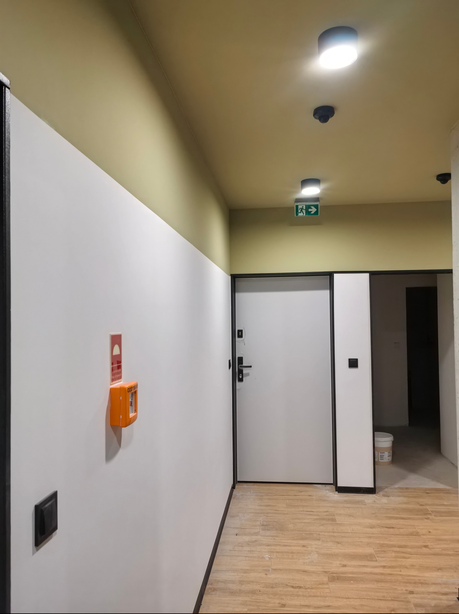 Korytarz po remoncie: białe ściany, drewnopodobne panele, żółty sufit z oświetleniem. Widoczny alarm przeciwpożarowy i drzwi z czarną ramą.