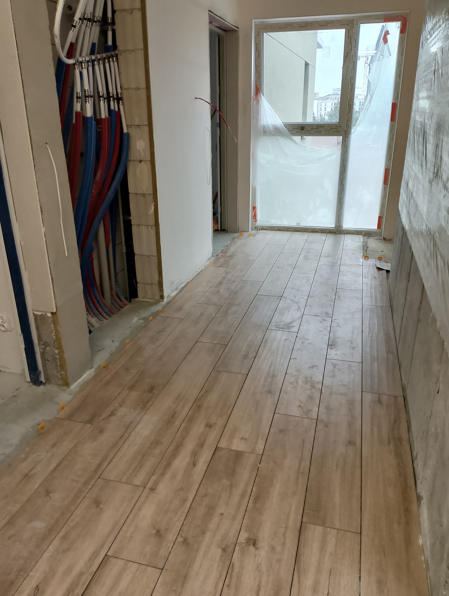 Układanie podłogi z płytek imitujących drewno w korytarzu. Widoczne rury instalacyjne przy ścianie i okno z folią ochronną. Użyto pomarańczowych krzyżyków dystansowych.