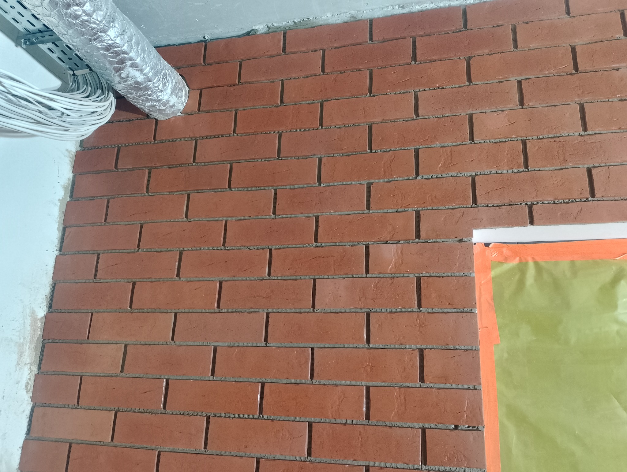 Ściana wykończona czerwoną cegłą dekoracyjną z widoczną instalacją wentylacyjną i elektryczną pod sufitem. Fragment okna zabezpieczony folią malarską.