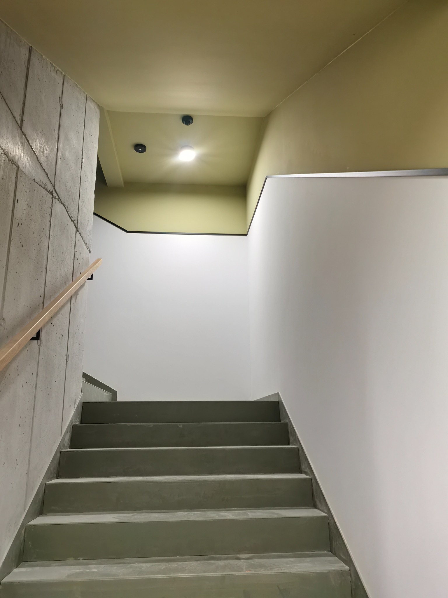 Betonowe schody z drewnianą poręczą w nowoczesnym wnętrzu. Ściany pomalowane na biało, sufit w odcieniach żółci. Widoczne oświetlenie sufitowe i czujniki.