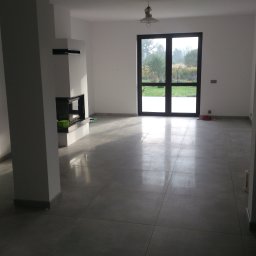 Art.Profil - Jasne, przestronne wnętrze po remoncie w Sosnowcu. Szare płytki podłogowe, białe ściany, nowoczesny kominek i duże okno z widokiem na zieleń.