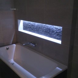 Art.Profil - Nowoczesna łazienka z wanną obudowaną ciemnymi płytkami i wnęką z dekoracyjną mozaiką podświetloną ledami, wykończona w szarościach.