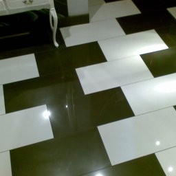 Art.Profil - Ukończona podłoga z błyszczących, prostokątnych płytek w szachownicę (biel i ciemny brąz), widoczny fragment białej komody na nóżkach.